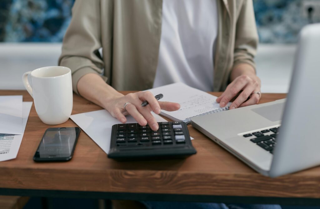 A person using a calculator and laptop while taking notes at a wooden desk.